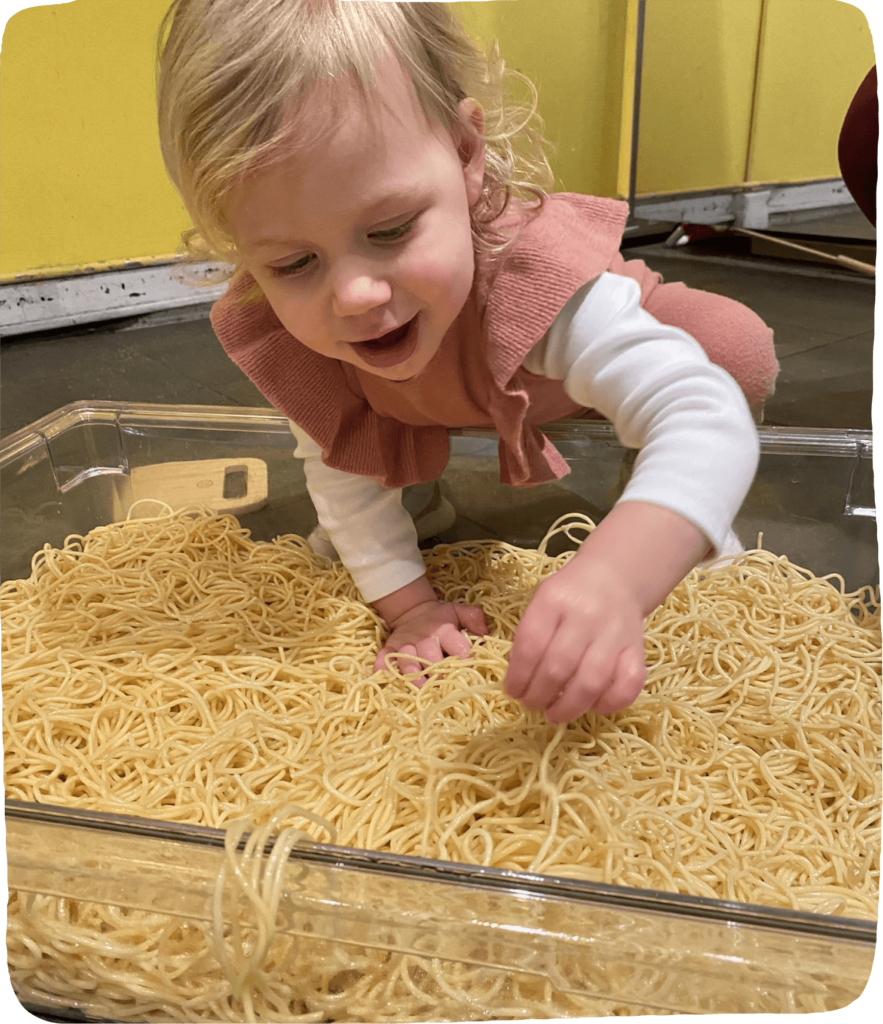 small girl reaching into a big bin of wet spaghetti