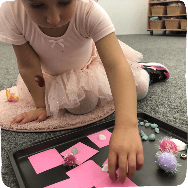 Image of a young child engaged in a play-based learning activity at home using loose parts such as pebbles, pink sticky notes, and pompoms.