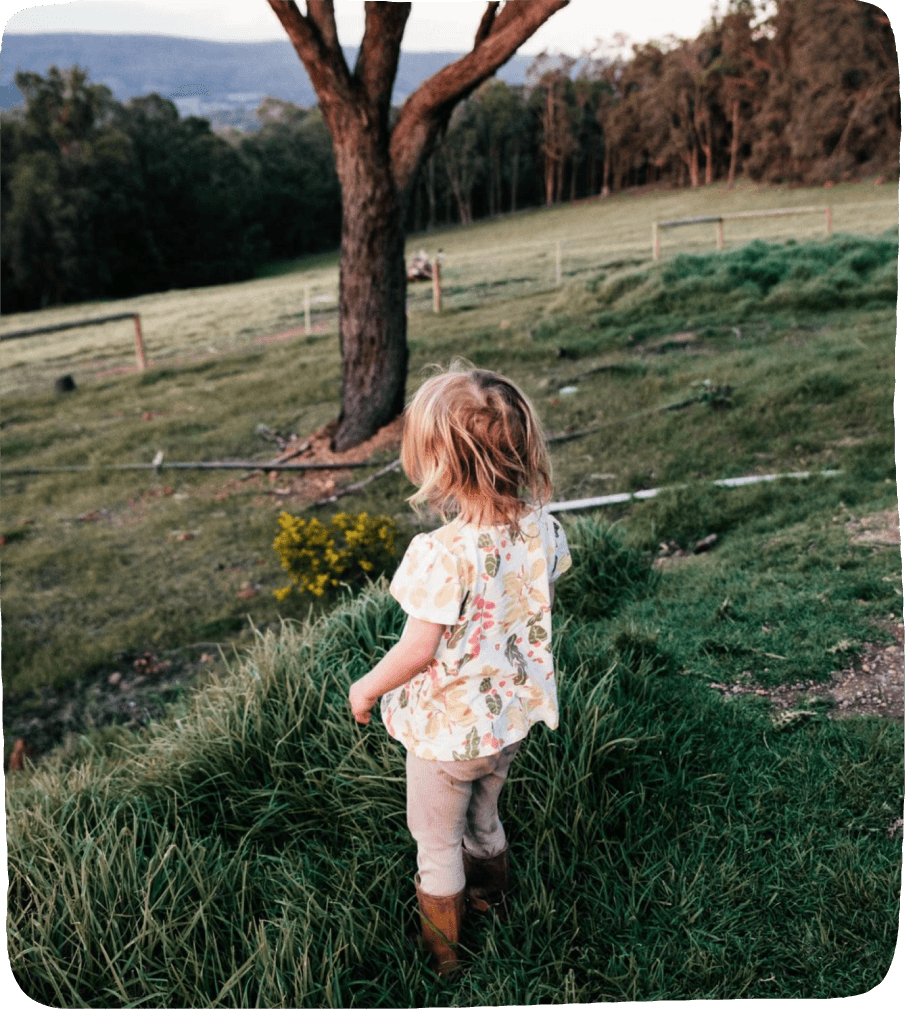 Image of a small girl seen from behind, walking through the grass near a tree to find the perfect spot for a summer nature activity.