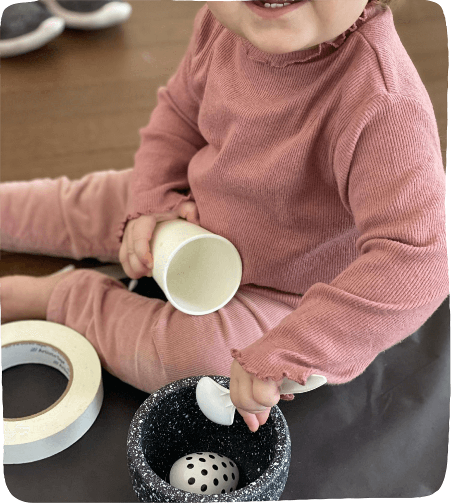 Image of a child engaging in a play-based learning activity using black & white materials such as a bowl, spoon, roll of masking tape, plastic cup, and a musical egg shaker. 