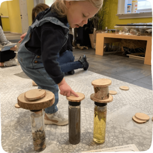 Child pointing (as if counting) to three sensory bottles with wooden tree logs on top. 