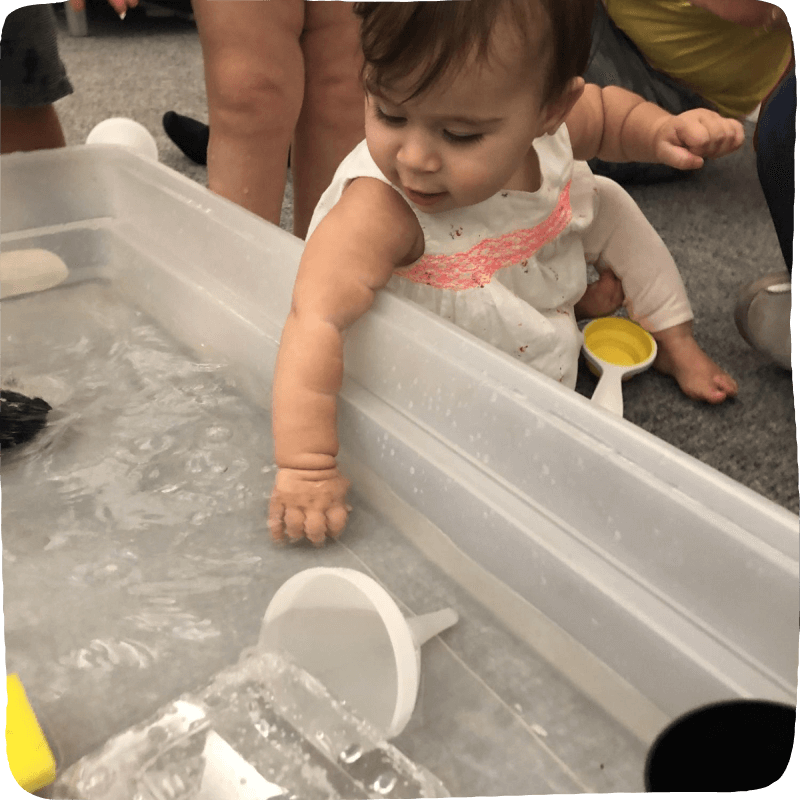 child reaching into a large tub of water to explore. Water play tools such as funnels and sponges are in the tub, ready to be played with.