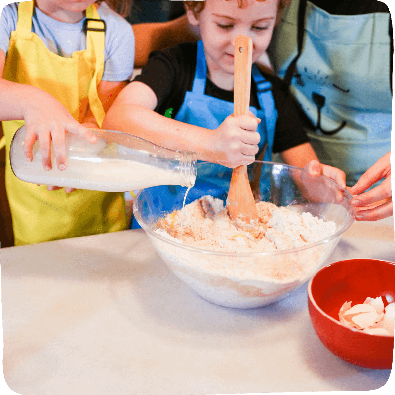 One child mixing flour and eggs in a bowl while another child adds in milk from a jar. This is a perfect example of how math happens when you cook with your children!