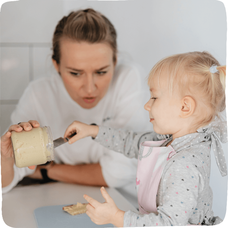 Adult holds a jar for child to use a knife to scoop and spread its contents on bread.