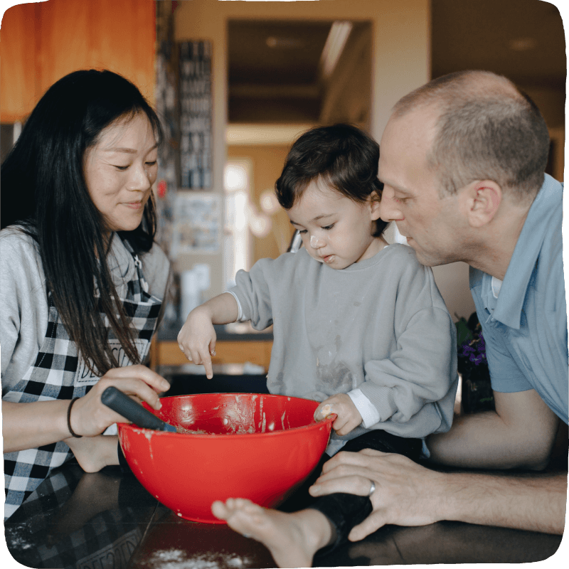 Two adults and a child doing a cooking activity together. They are experiencing math as it occurs while looking into a large red mixing bowl as the child points to its contents. 