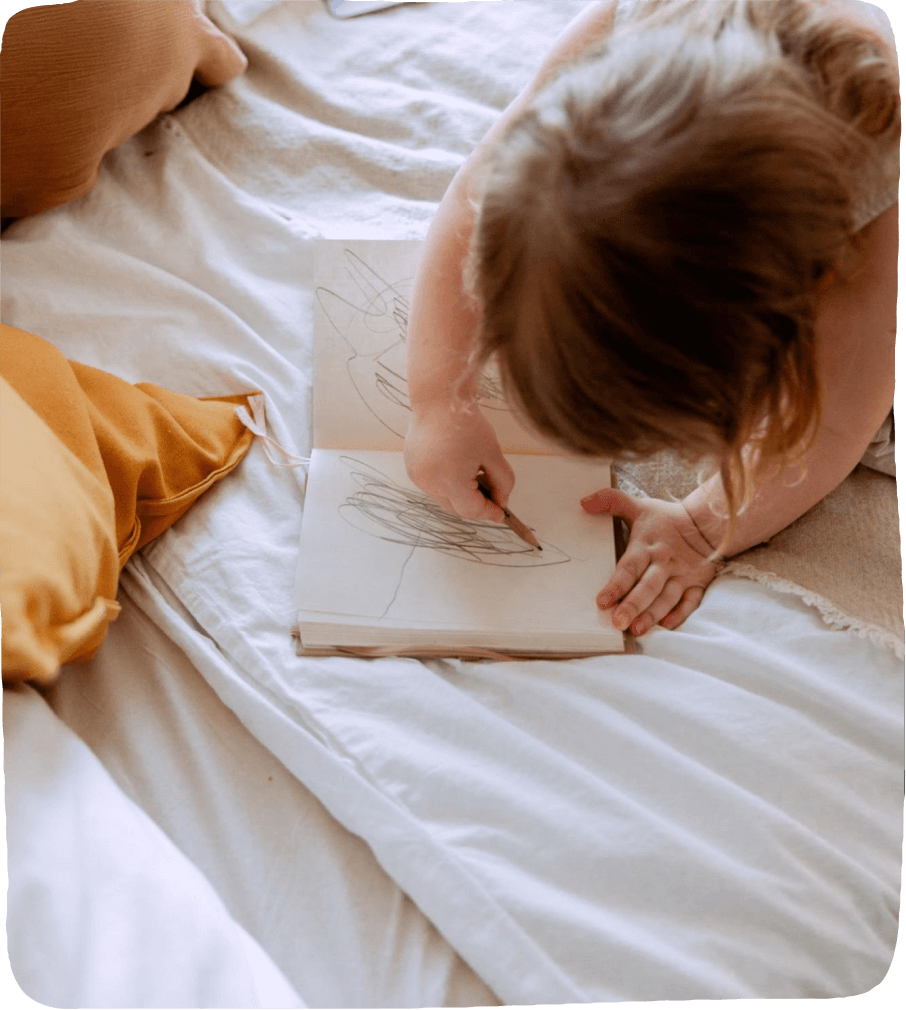 A child lays on a bed and uses a single drawing utensil to create art in her journal