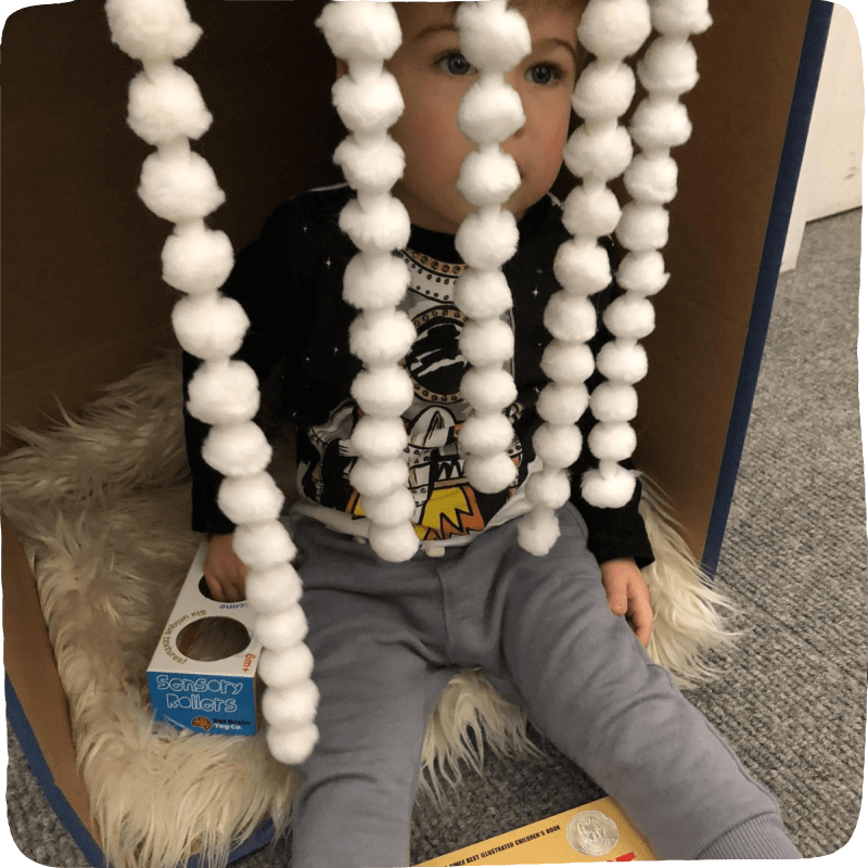 Child sits in a box with a cozy rug and hanging soft white cotton balls. 