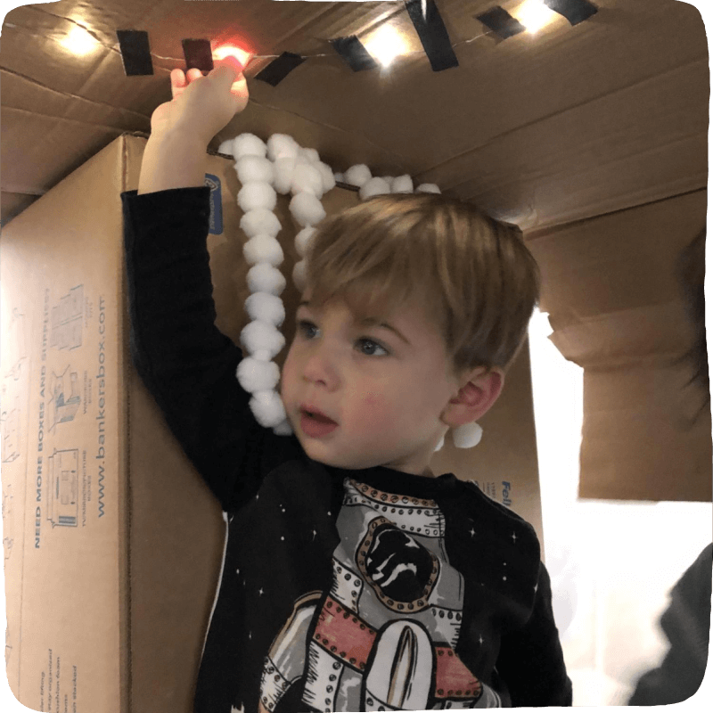 Child sitting in a box fort. White cottons balls and Italian lights have been hung on the box top to create a cozy warm nook. 