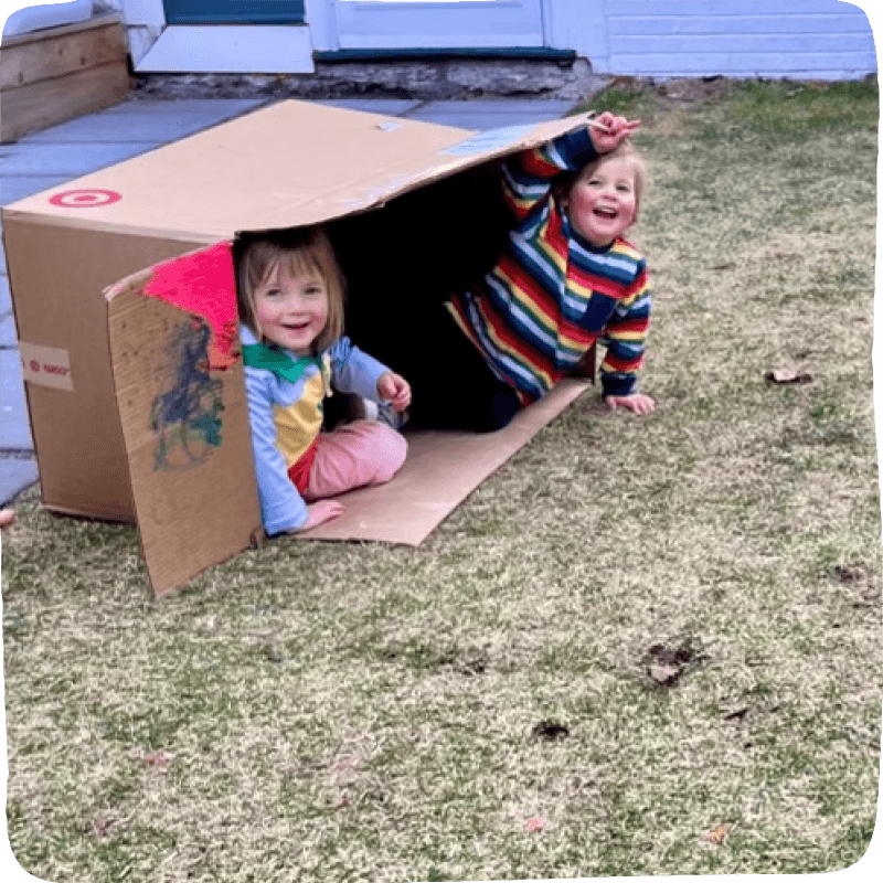 Two kids in the yard smiling and playing in a cardboard box.