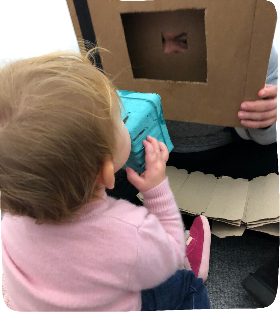 Toddler girl looking through a cardboard berry container through a cardboard box with a hole in it to illustrate The Value of cardboard box Play.