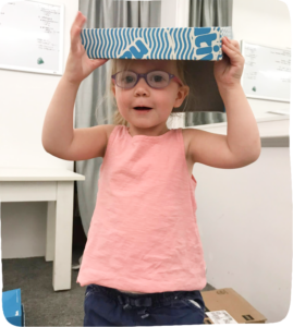 Little girl with a shoe box on her head to highlight The Value of Cardboard Box Play.