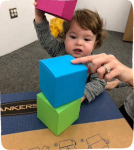 Little toddler stacking colorful cardboard boxes and a mother's hand pointing.